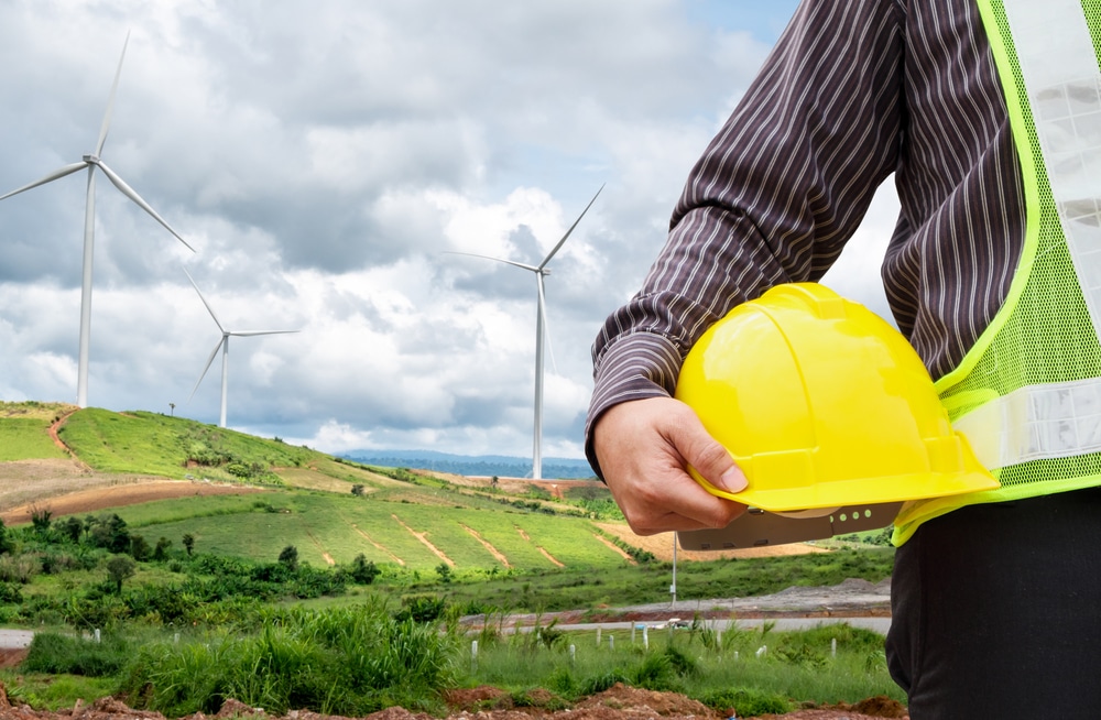 Close-up of man wearing yellow vest and carrying yellow hat, wind turbines in background